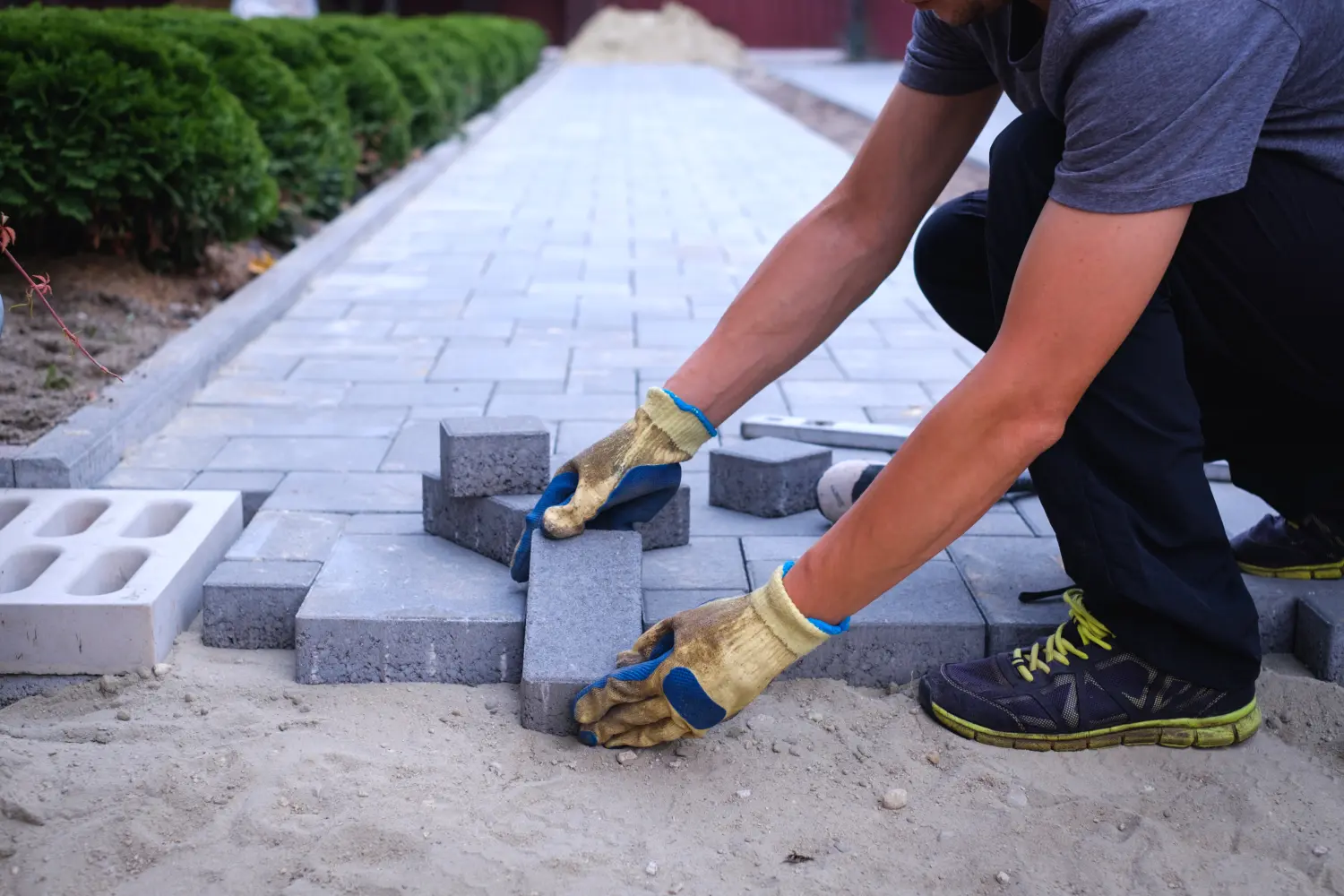 Der Meister mit den gelben Handschuhen verlegt schichtweise Pflastersteine. Pflasterung eines Gartenwegs mit Ziegelsteinen durch einen professionellen Pflasterer.